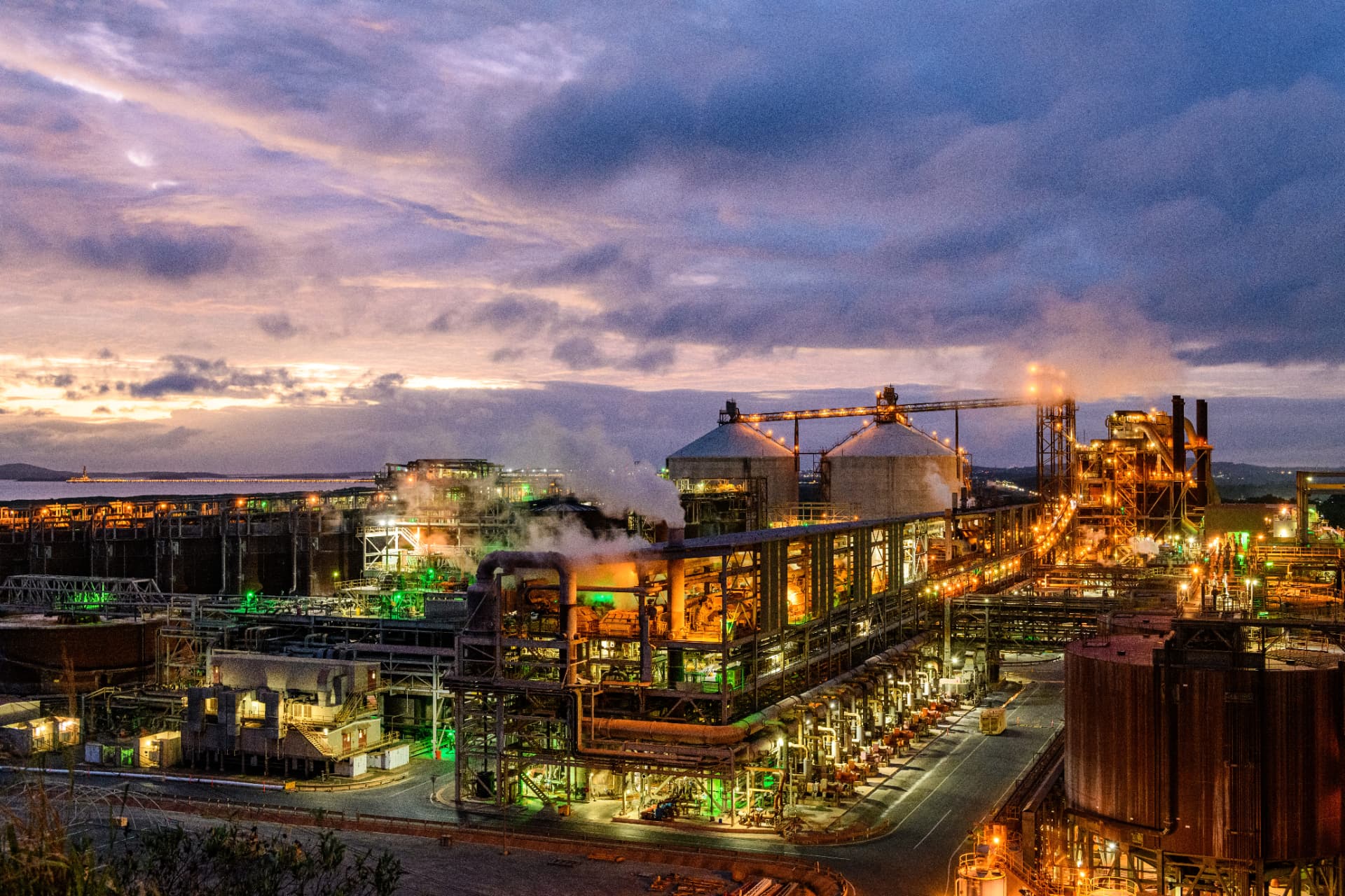 Industrial processing facility at twilight, lit process trains, vessels, stacks and steam against a coloured sky